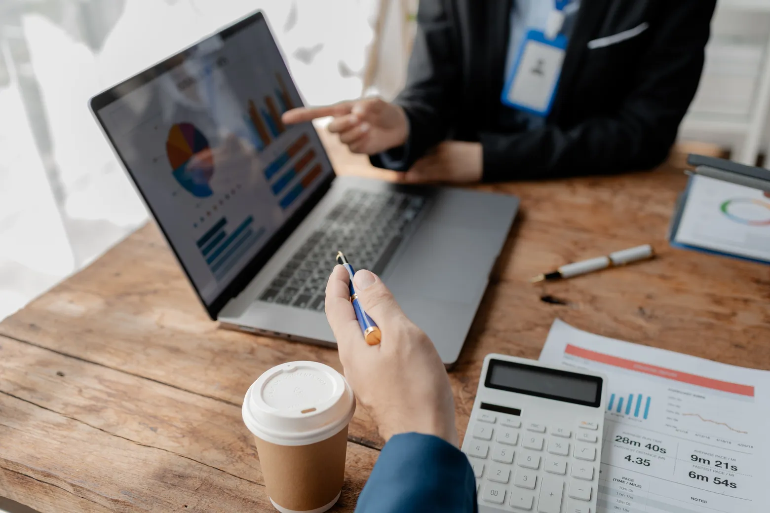 To personer analyserer data på en bærbar datamaskin med diagrammer og grafer. Den ene peker på skjermen, og den andre holder en penn. En kalkulator, papirrapporter og en kaffekopp står på trebordet.