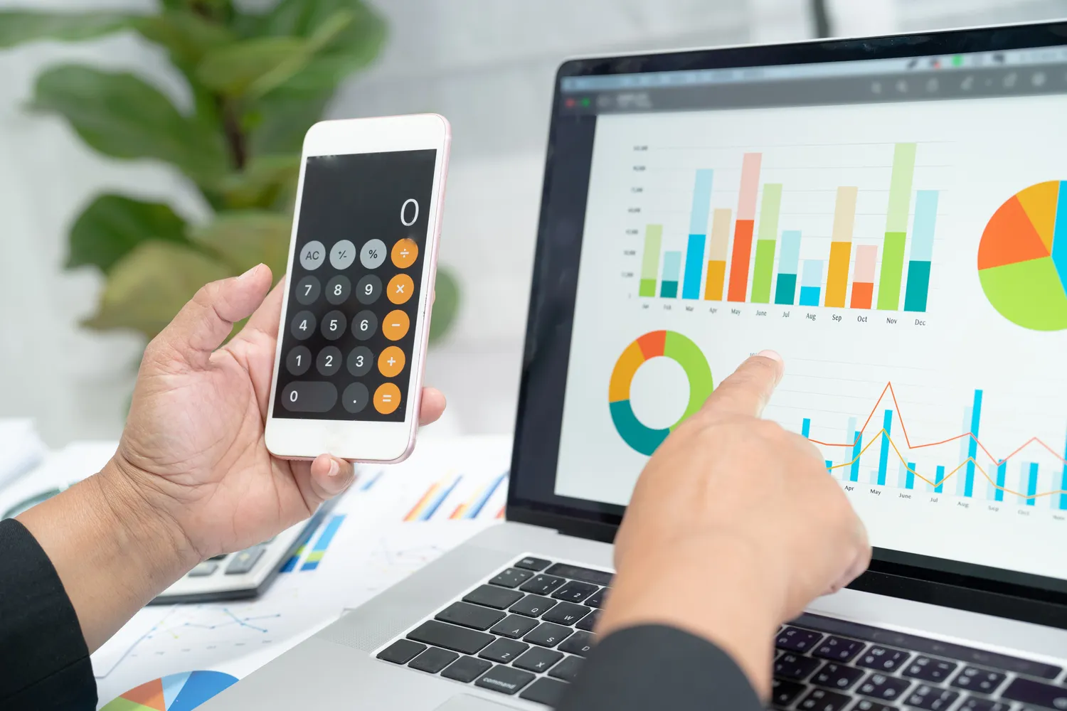 En person holder en smarttelefon som viser en kalkulator-app mens han peker på grafer på en bærbar datamaskin. Grafene inneholder søylediagrammer og sektordiagrammer, som sannsynligvis illustrerer økonomiske data. En plante er i bakgrunnen.