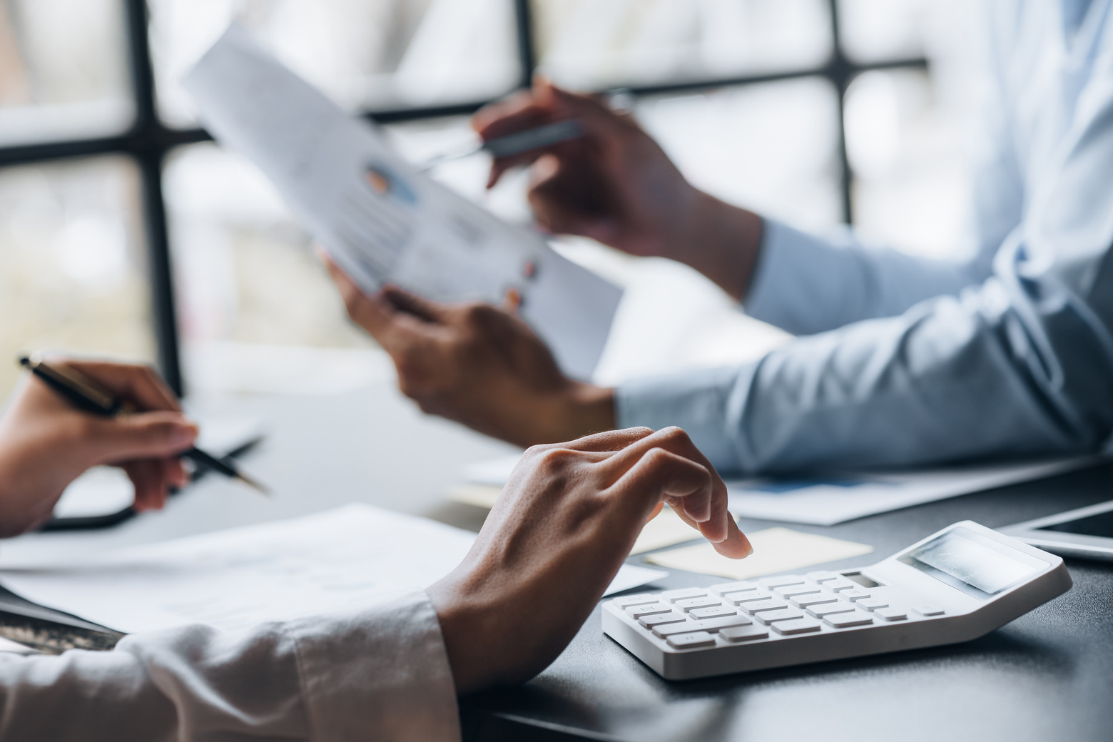 Et nærbilde av to hender som holder dokumenter og penner, med en kalkulator i forgrunnen. Personene er engasjert i en diskusjon, muligens analyserer data eller finansiell informasjon, i et sterkt opplyst kontormiljø.